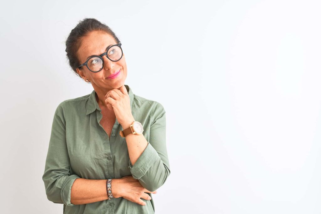 Middle age woman wearing green shirt and glasses standing over isolated white background with hand on chin thinking about question, pensive expression. Smiling with thoughtful face. Doubt concept. - stock photo
