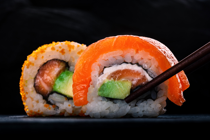 Two sushi rolls with salmon and avocado, on a black background with chopsticks