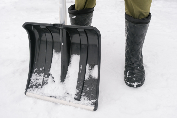 An unrecognizable person in snow boots holding a plastic shovel in order to clean, remove snow from a sidewalk for safety