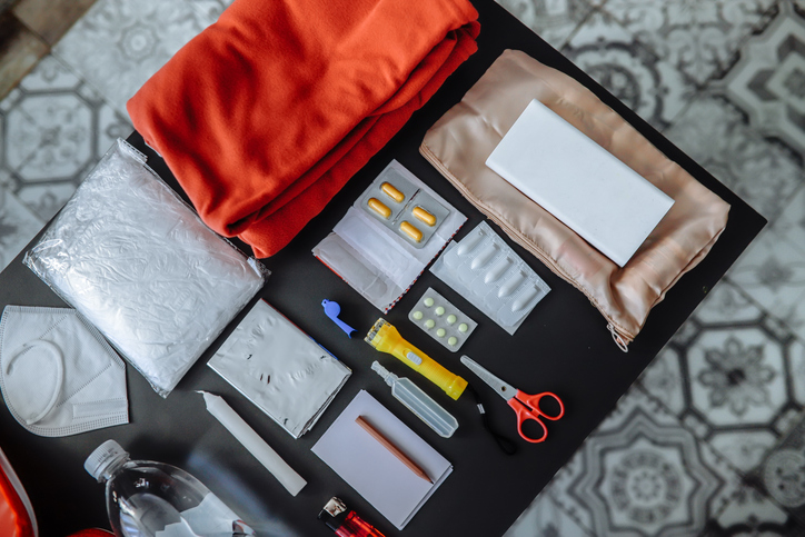 Prepping flat lay: survival gear including water bottle, raincoat, power bank, and a notebook for emergency planning, organized in a real room.
