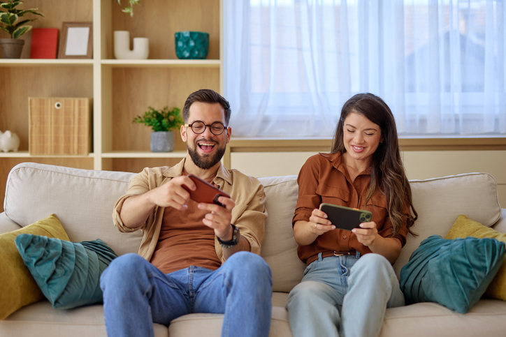 Young couple playing videogames on smartphones while sitting on sofa at home