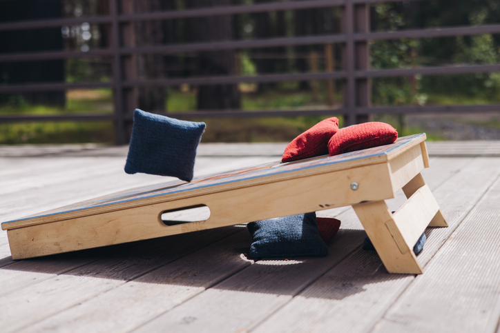 Cornhole game set, process of throwing bean bags, female girl tossing bean sacks, corn hole in the backyard, wooden boards for corn-hole tournament in a summer day on a wooden flooring terrace deck