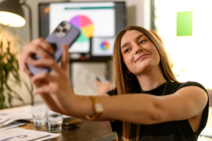 Young woman takes a selfie while sitting at her desk in a bright modern office during a creative brainstorming session with coworkers nearby